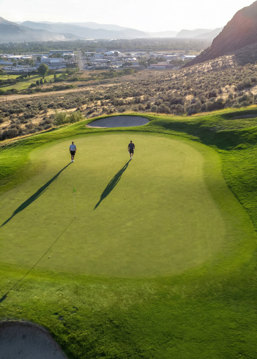 Aerial view of two golfers walking across a sunlit green at Bighorn Golf Course overlooking Kamloops valley
