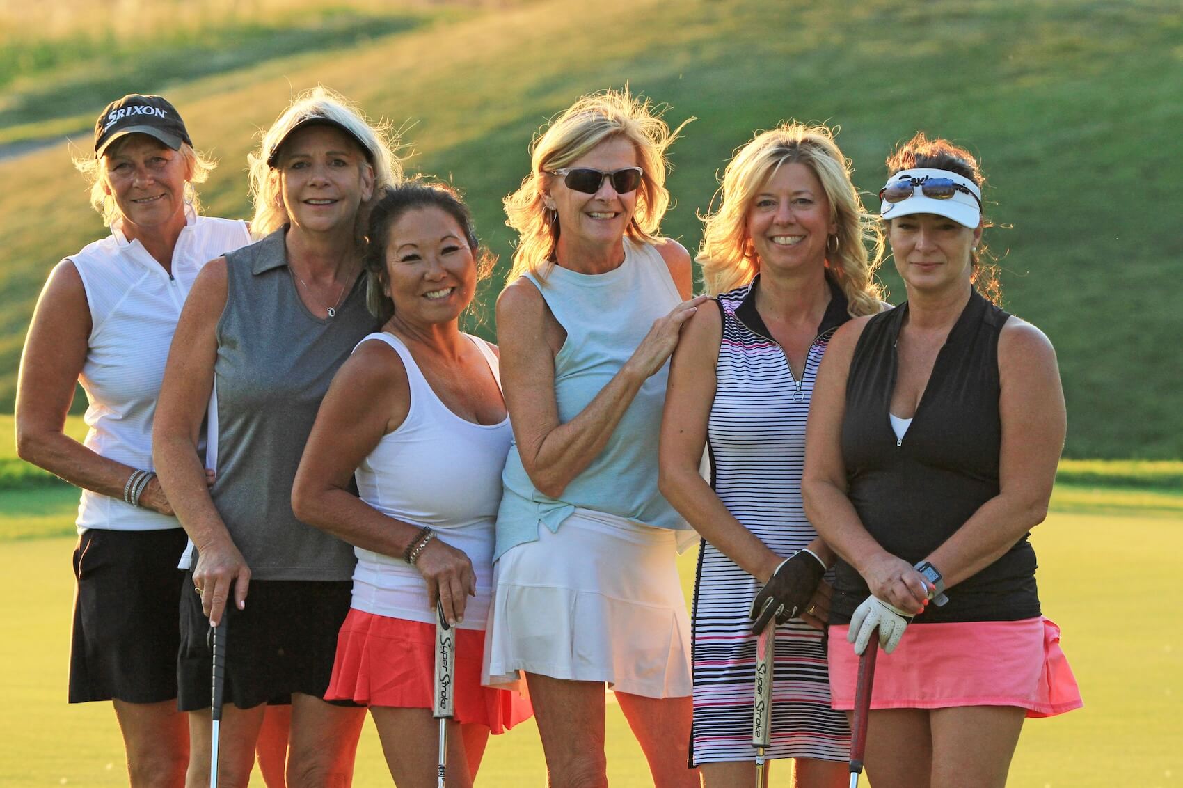 A group of six smiling women in golf attire pose together on a green at Bighorn Golf & Country Club during a Ladies Day event, with the soft glow of the late afternoon sun in the background.