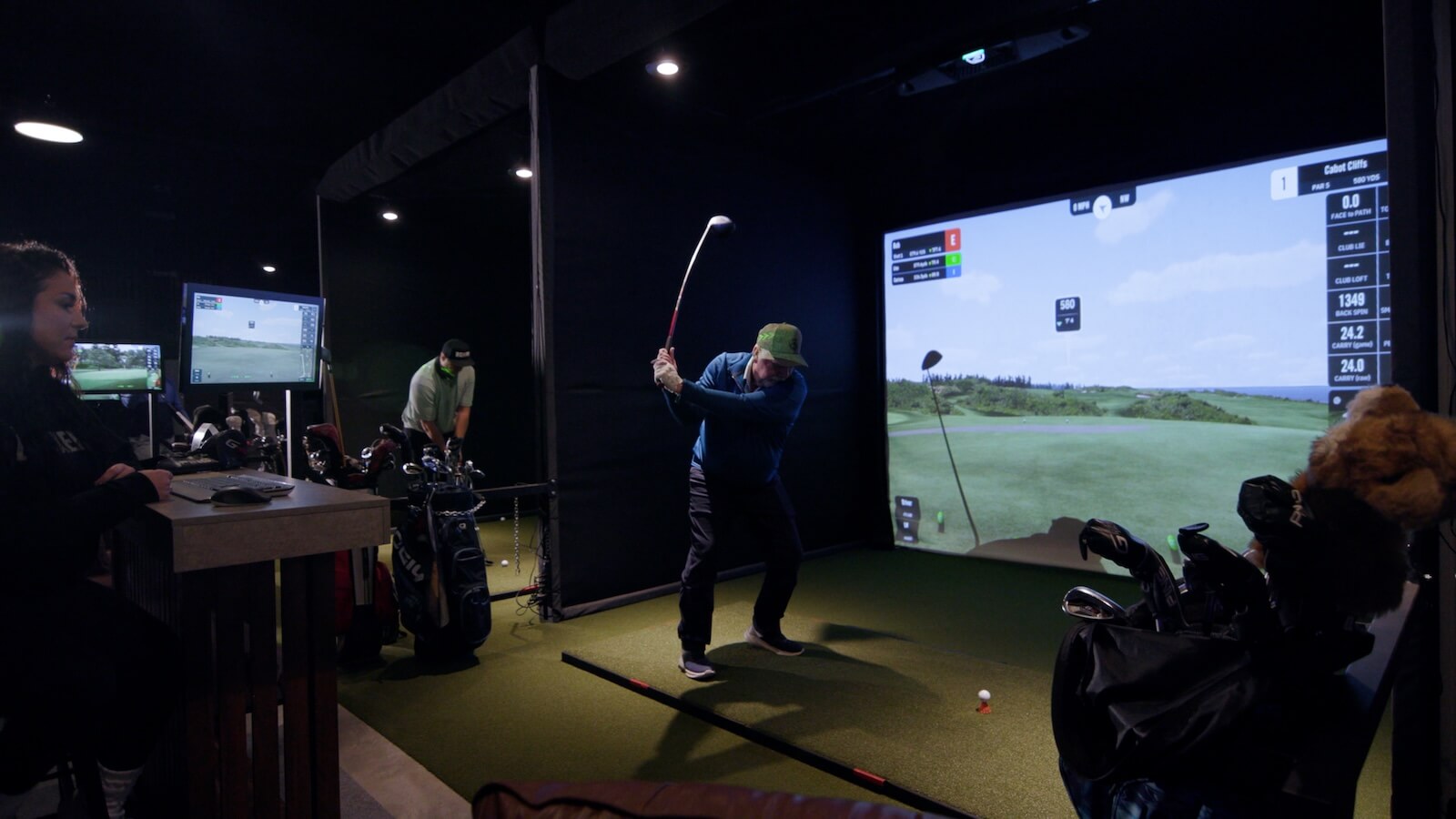Golfer mid-swing in an indoor golf simulator bay with virtual course projection and equipment setup